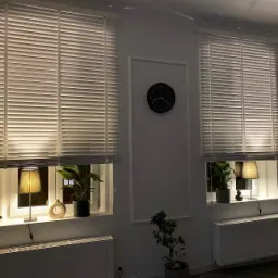 Dwa okna z białymi żaluzjami aluminiowymi, oświetlone lampkami, z zegarem na ścianie pomiędzy nimi.
