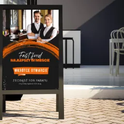 Czarny, metalowy stojak reklamowy z plakatem informującym o otwarciu restauracji Fast Food. Na plakacie uśmiechnięta para kelnerów w tle restauracji oraz napis 'Fast Food NAJLEPSZY W MIEŚCIE'...