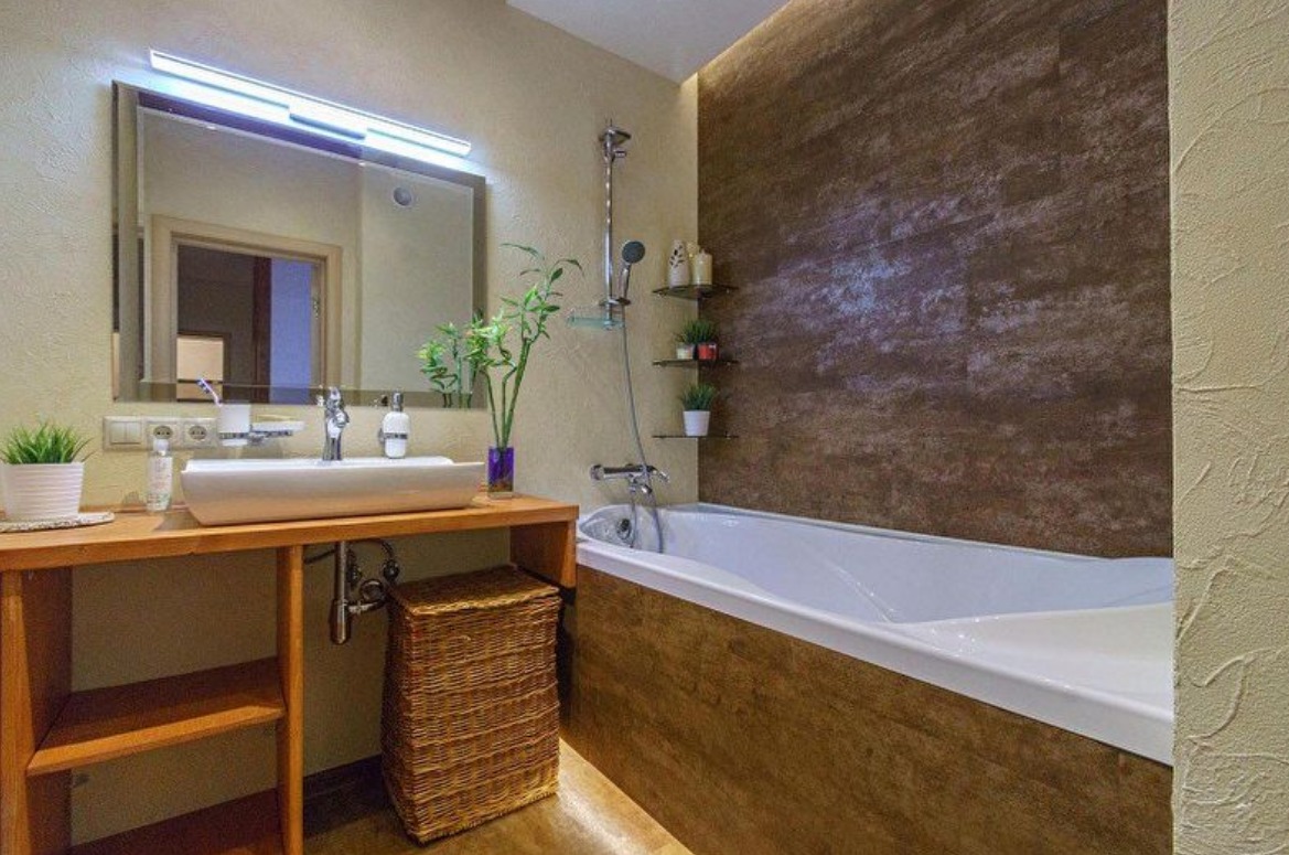 Nowoczesna łazienka z drewnianymi akcentami: umywalka nablatowa, wanna obudowana drewnem, ściana z teksturą, lustro z oświetleniem LED i wiklinowy kosz.