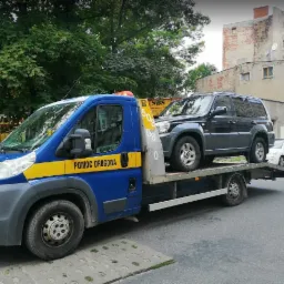 Laweta pomocy drogowej przewozi czarnego SUV-a na tle miejskiej zabudowy.