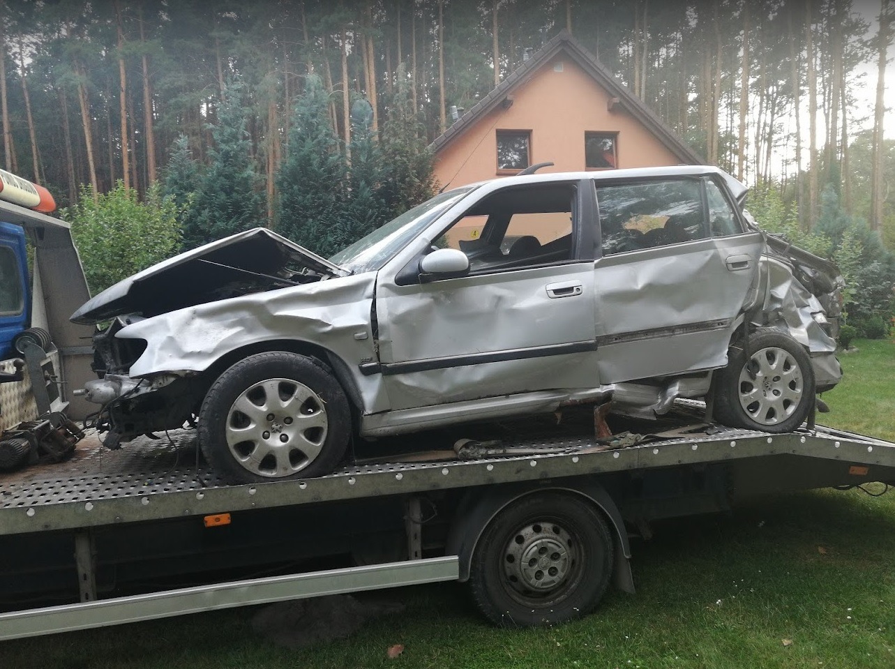 Srebrny samochód osobowy po wypadku, z poważnymi uszkodzeniami karoserii, załadowany na lawetę holowniczą na tle domu i drzew.