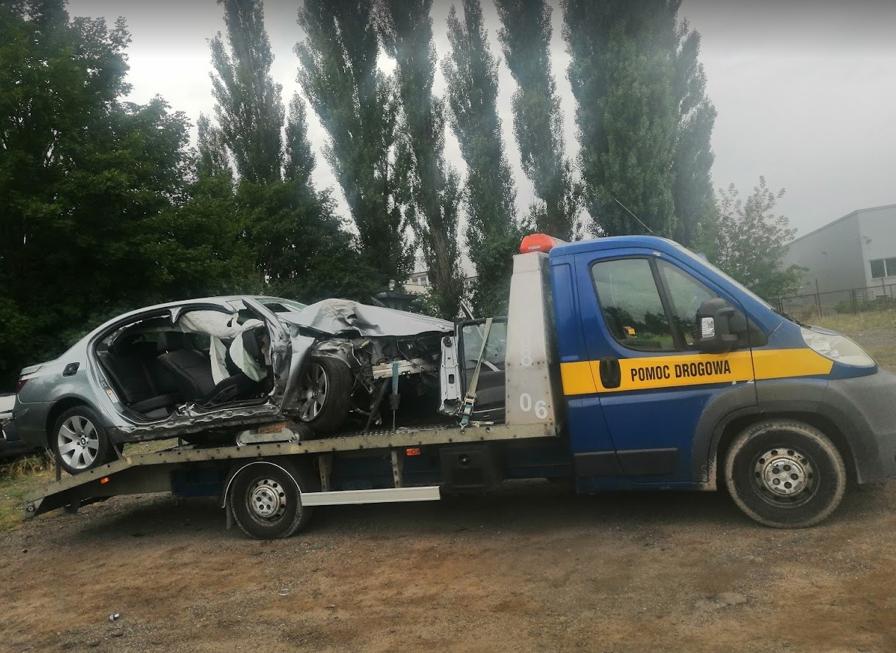 Uszkodzony srebrny samochód BMW na lawecie pomocy drogowej z widocznymi wystrzelonymi poduszkami powietrznymi, na tle drzew i szarego nieba.