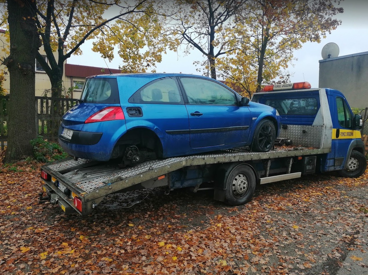 Niebieski samochód bez jednego koła załadowany na lawetę pomocy drogowej na tle jesiennych liści.