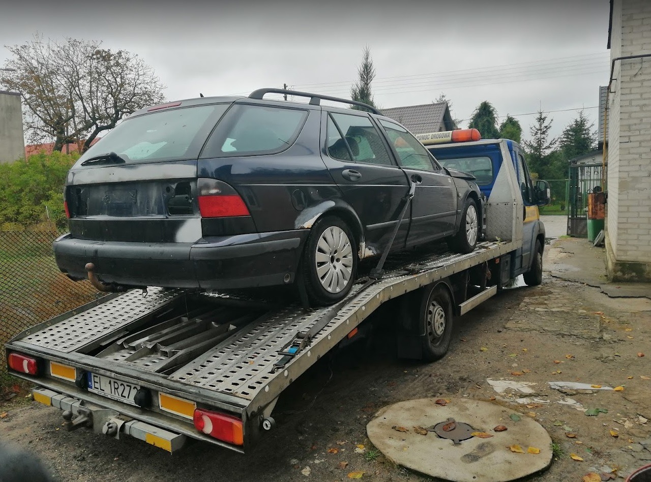 Czarny Saab kombi bez tablic rejestracyjnych, z widocznymi uszkodzeniami, transportowany na lawecie pomocy drogowej.