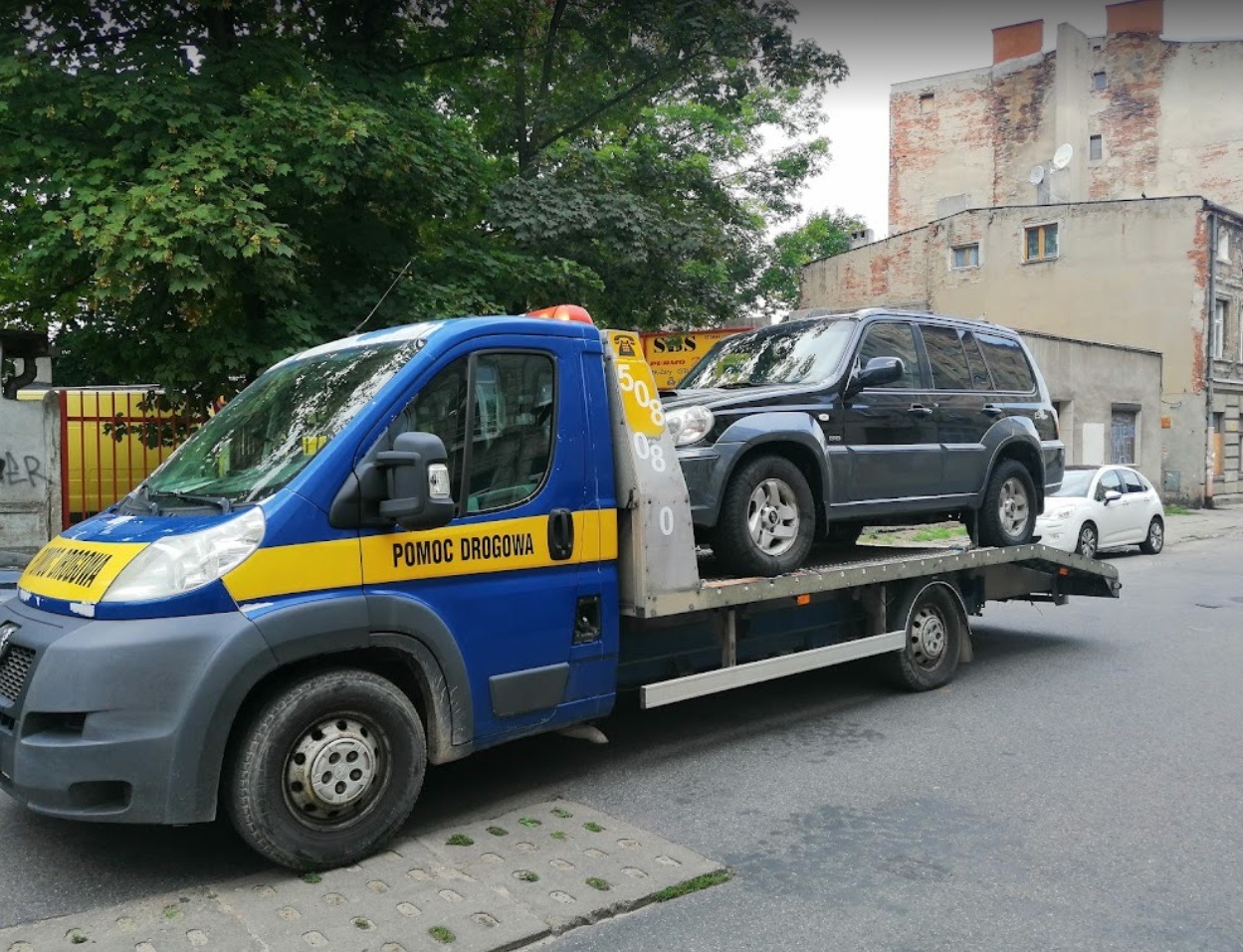 Laweta pomocy drogowej przewozi czarnego SUV-a na tle miejskiej zabudowy.