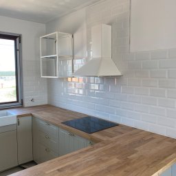 HD wykończenia wnetrz - Nowoczesna kuchnia z białymi, błyszczącymi płytkami typu metro na ścianie, drewnianym blatem, szarymi szafkami z złotymi uchwytami i czarną płytą indukcyjną. Widoczny biały okap i półka.