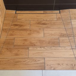 HD wykończenia wnetrz - Fragment wykończonej łazienki z brązowym, kafelkowym sufitem, drewnopodobnymi płytkami na ścianie i podłodze oraz jasnymi, kwadratowymi płytkami podłogowymi.