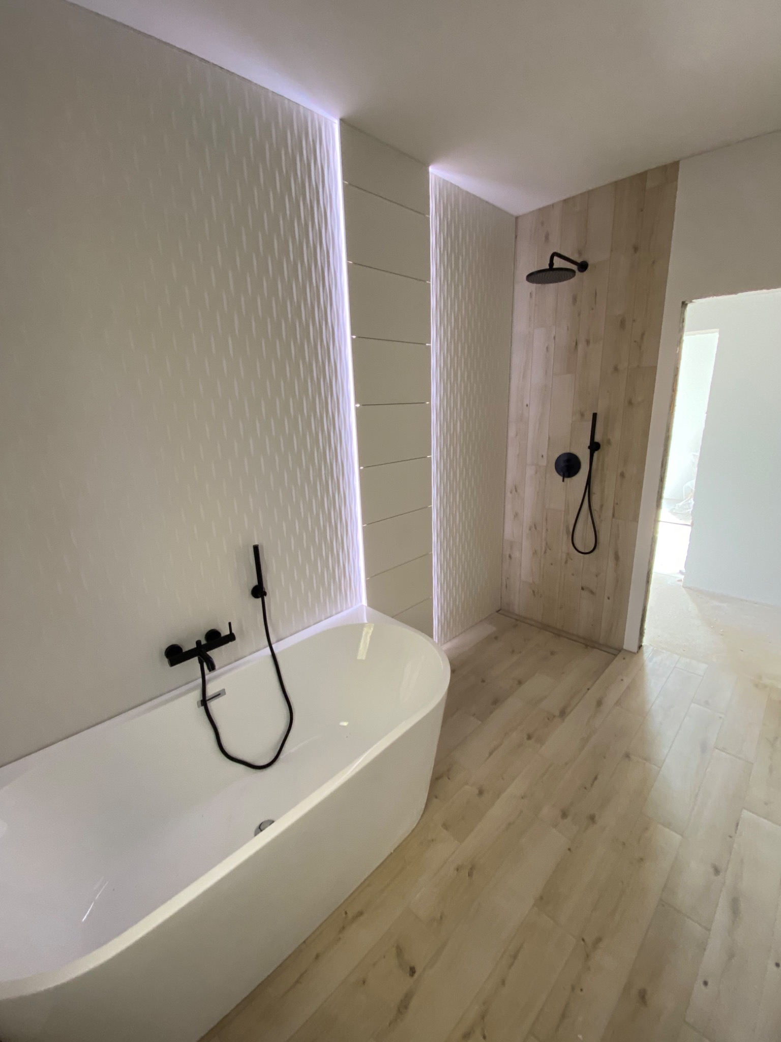 Nowoczesna łazienka z wolnostojącą białą wanną, czarną armaturą i kabiną prysznicową wyłożoną panelami imitującymi drewno. Ściana z dekoracyjną fakturą i podświetleniem LED.