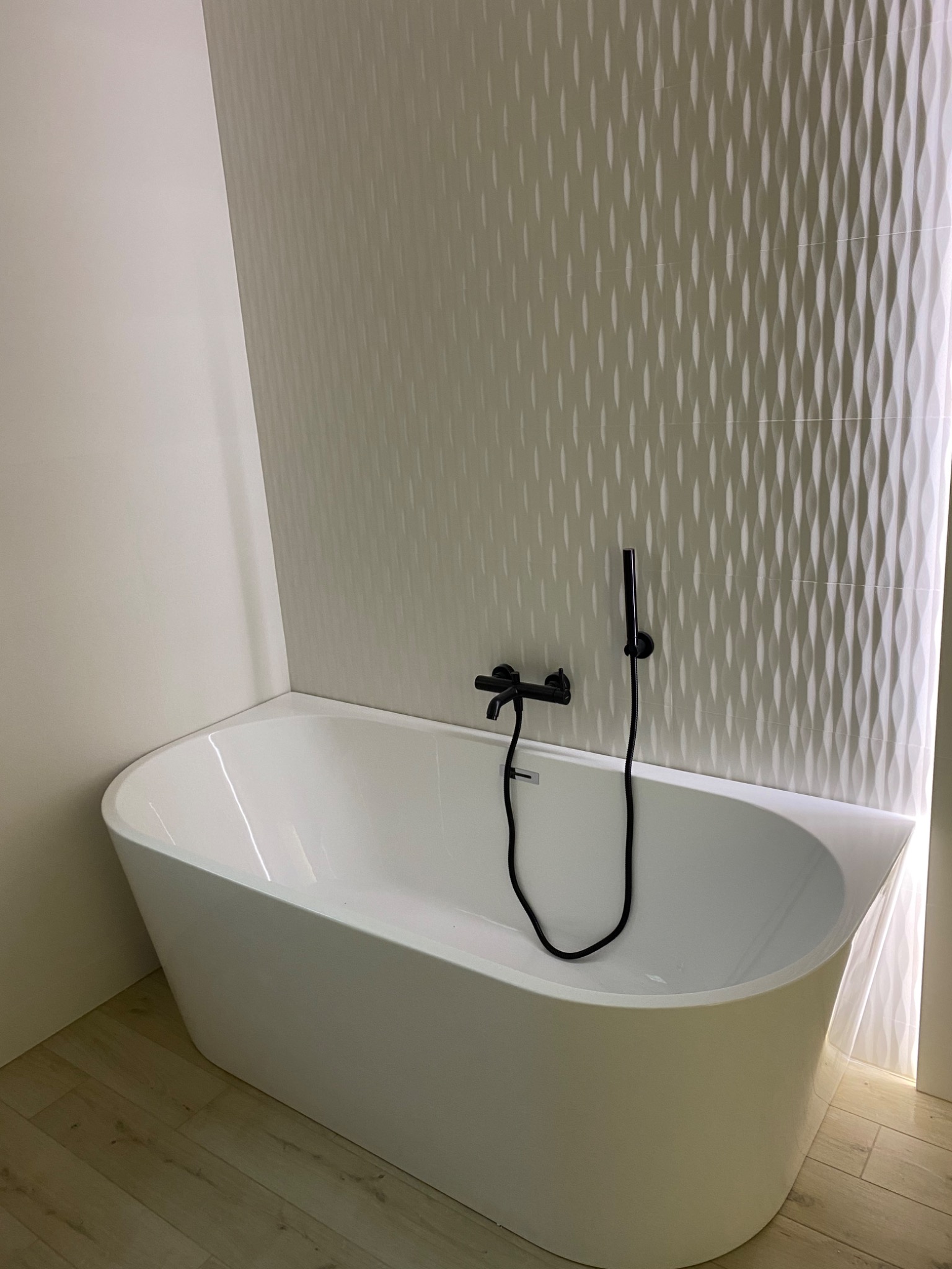 Nowoczesna biała wanna wolnostojąca z czarną armaturą na tle ściany z dekoracyjnymi, falistymi płytkami w odcieniach bieli i beżu.