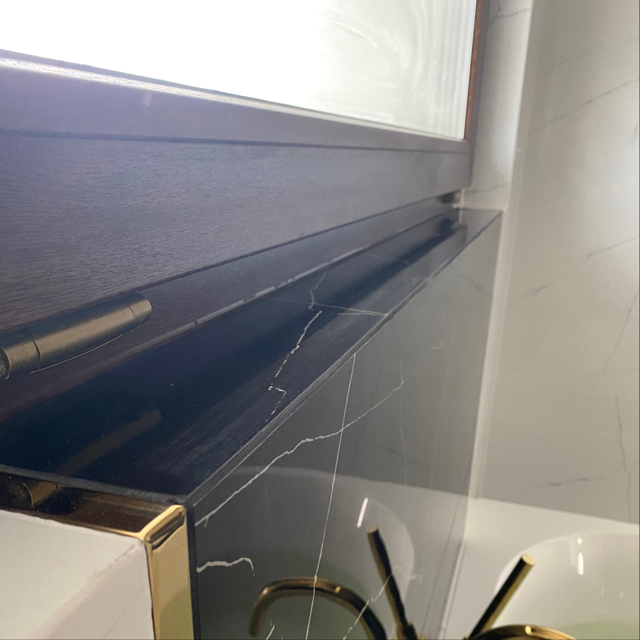 Szczegółowe ujęcie umywalki z czarnym blatem imitującym marmur z białymi żyłami i złotymi elementami wykończeniowymi, widoczne również fragmenty lustra i białych płytek na ścianie.
