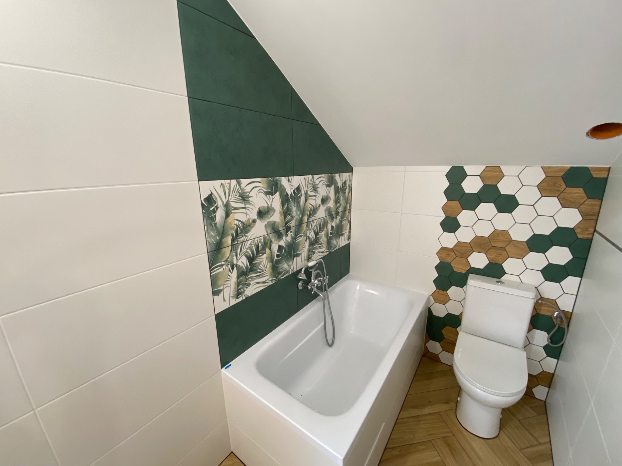 Wykończona łazienka w skosach z białą wanną, toaletą i mozaiką heksagonalną w odcieniach zieleni, bieli i drewna, oraz płytkami z motywem liści tropikalnych.