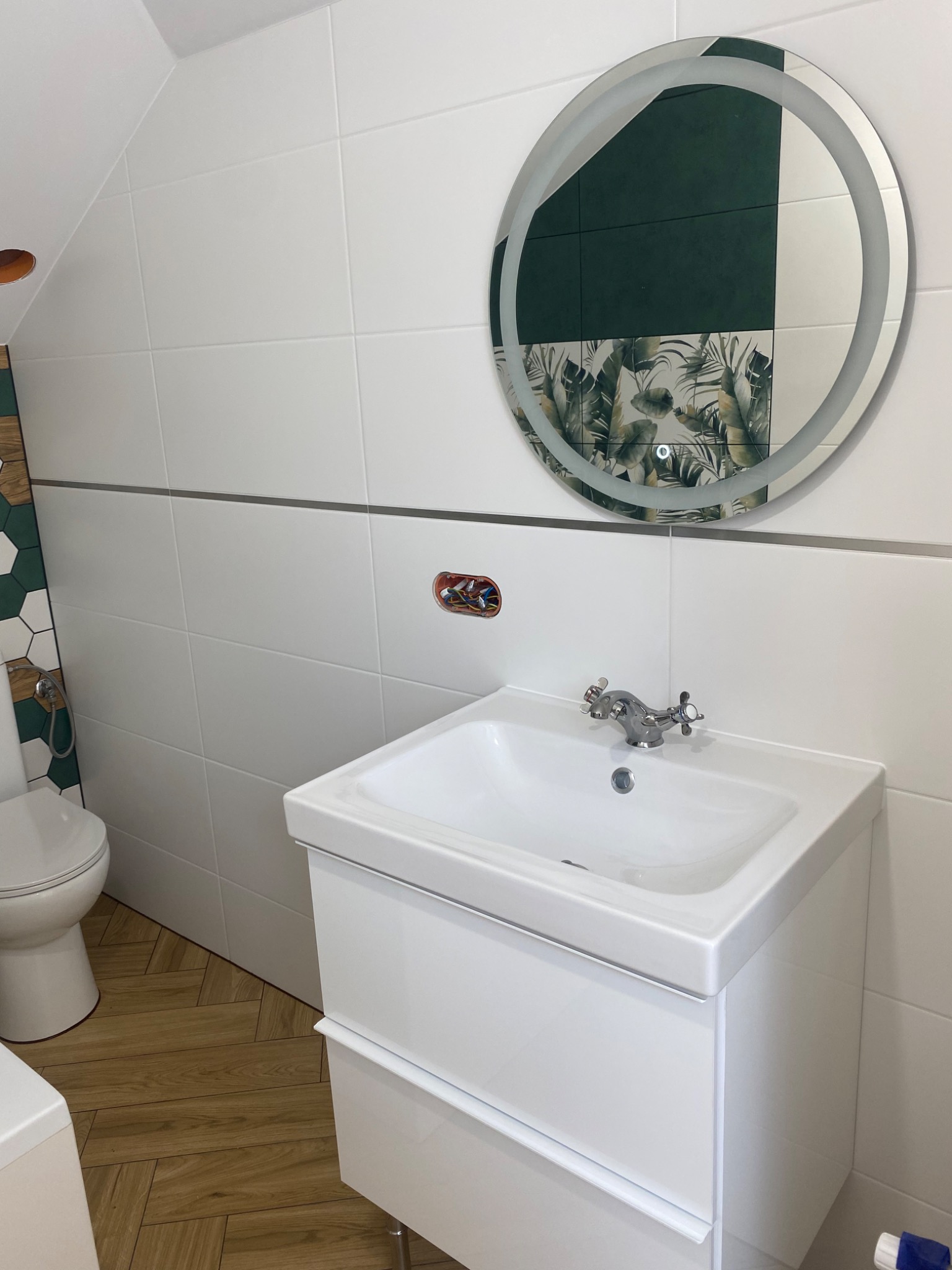 Jasna łazienka na poddaszu z drewnopodobną podłogą ułożoną w jodełkę, biała umywalka z szafką, okrągłe lustro z podświetleniem, białe płytki na ścianach z dekoracyjnym pasem i fragmentem ściany...