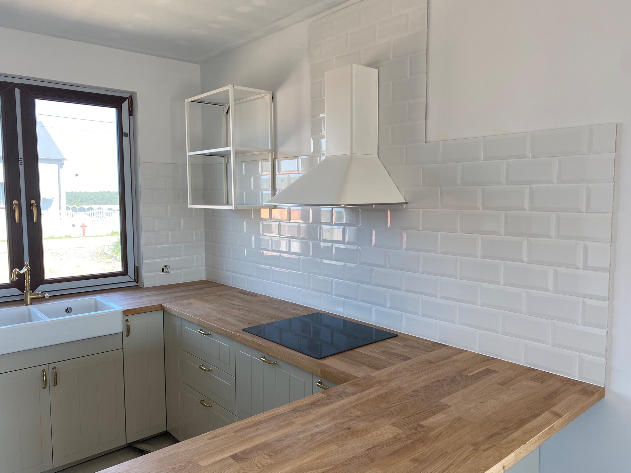 Nowoczesna kuchnia z białymi, błyszczącymi płytkami typu metro na ścianie, drewnianym blatem, szarymi szafkami z złotymi uchwytami i czarną płytą indukcyjną. Widoczny biały okap i półka.