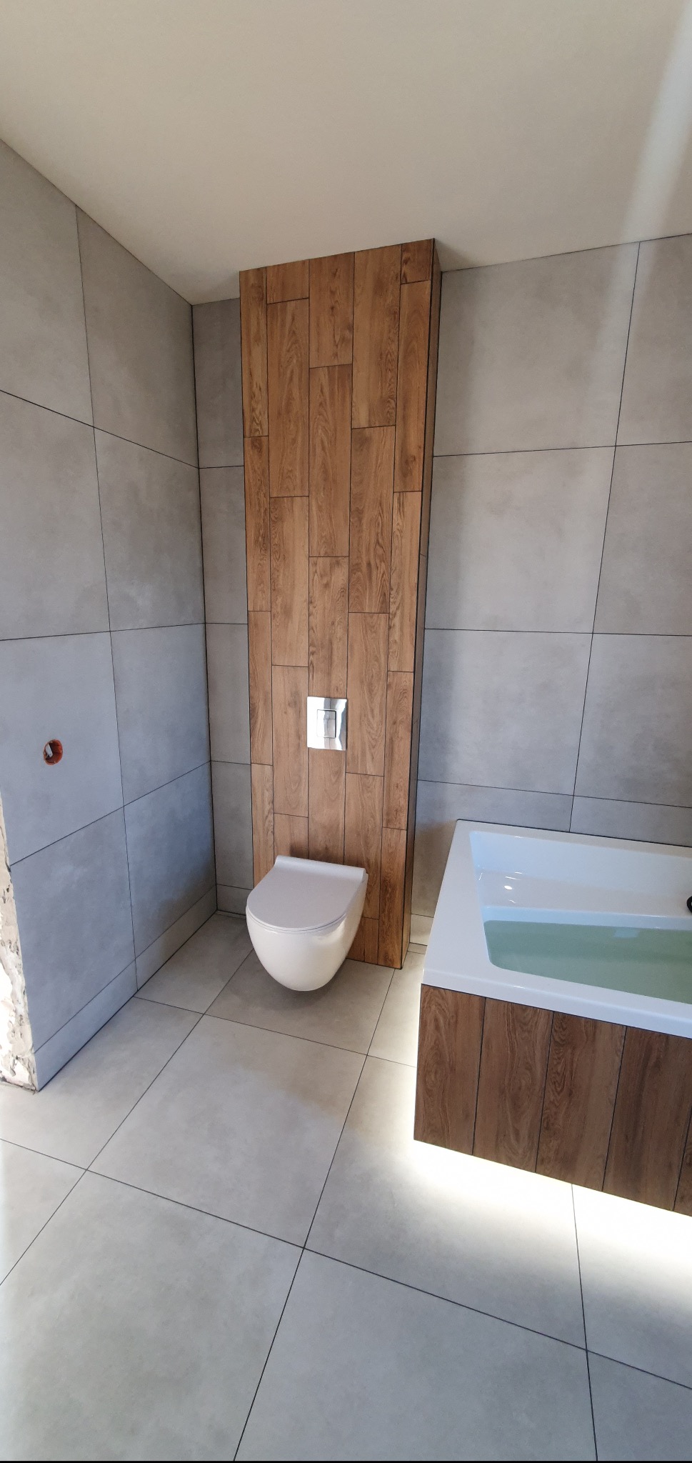 Wykończona łazienka z szarymi płytkami, drewnopodobną zabudową toalety i wanny z podświetleniem LED.