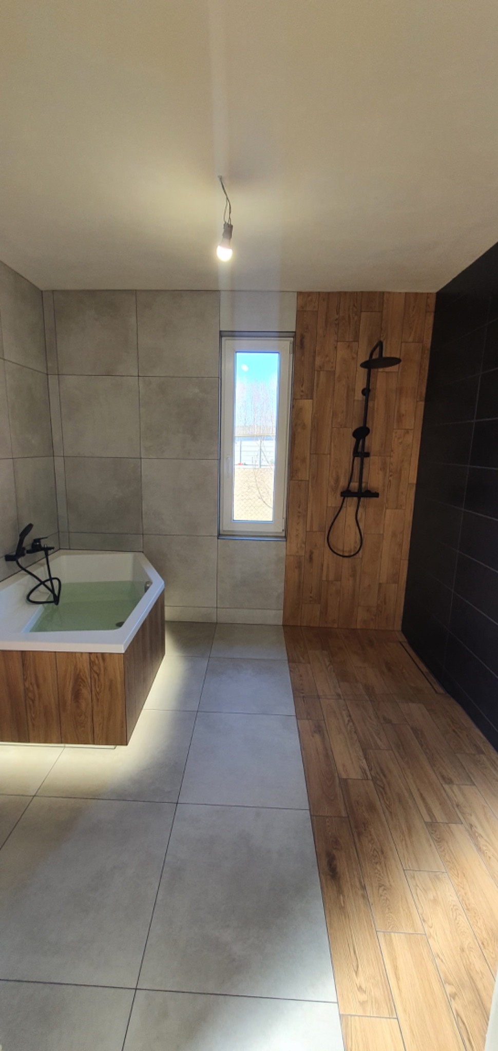Nowoczesna łazienka z wanną z podświetleniem LED, kabiną prysznicową typu walk-in z czarną armaturą i mieszanką płytek imitujących drewno i szarego betonu, oświetlona pojedynczą żarówką.