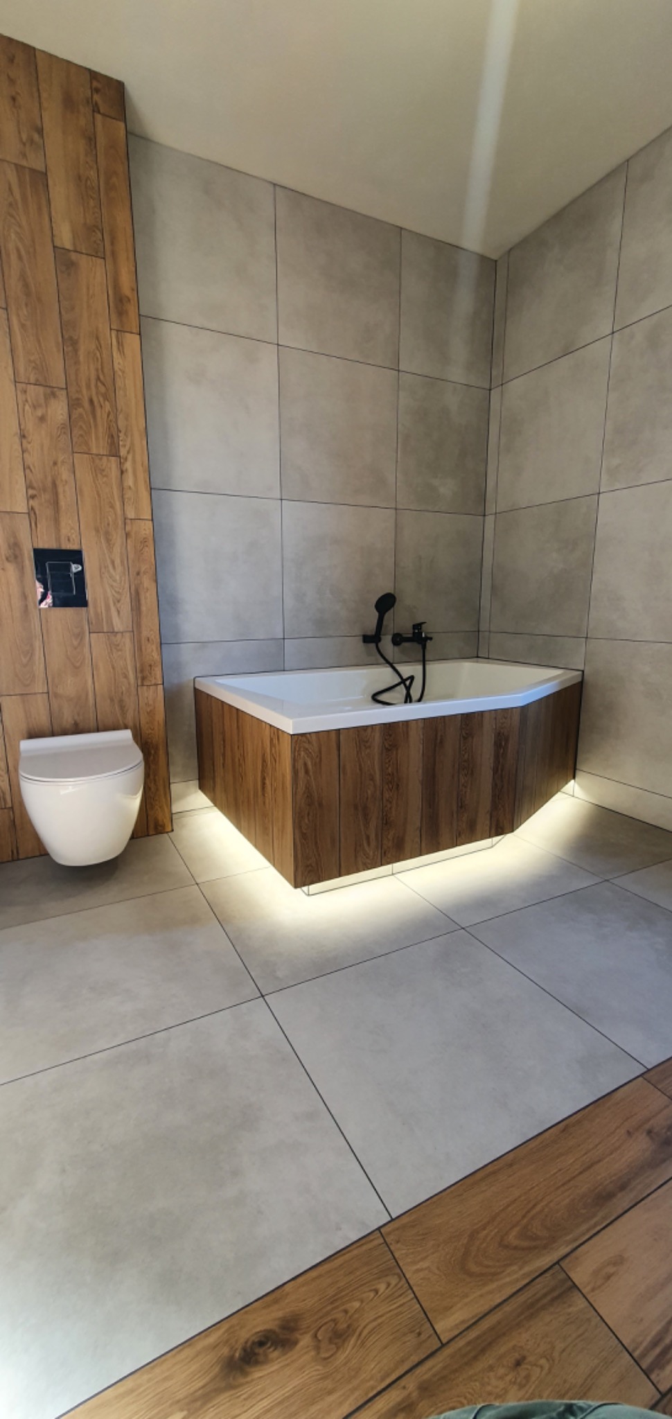 Nowoczesna łazienka z wanną obudowaną panelami imitującymi drewno, podświetloną od dołu, płytkami w odcieniach szarości na ścianach i podłodze oraz toaletą podwieszaną przy ścianie wykończonej...