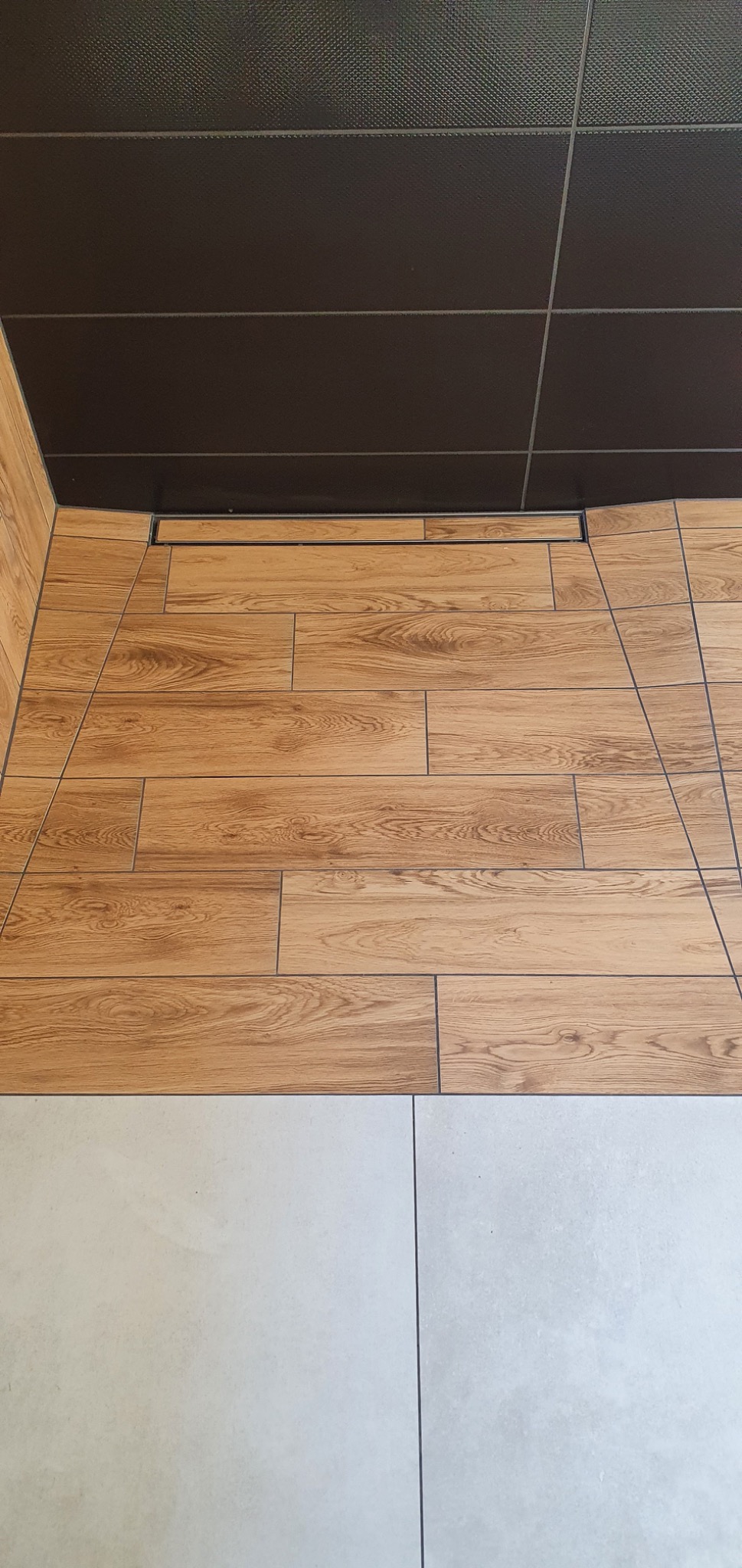 Fragment wykończonej łazienki z brązowym, kafelkowym sufitem, drewnopodobnymi płytkami na ścianie i podłodze oraz jasnymi, kwadratowymi płytkami podłogowymi.