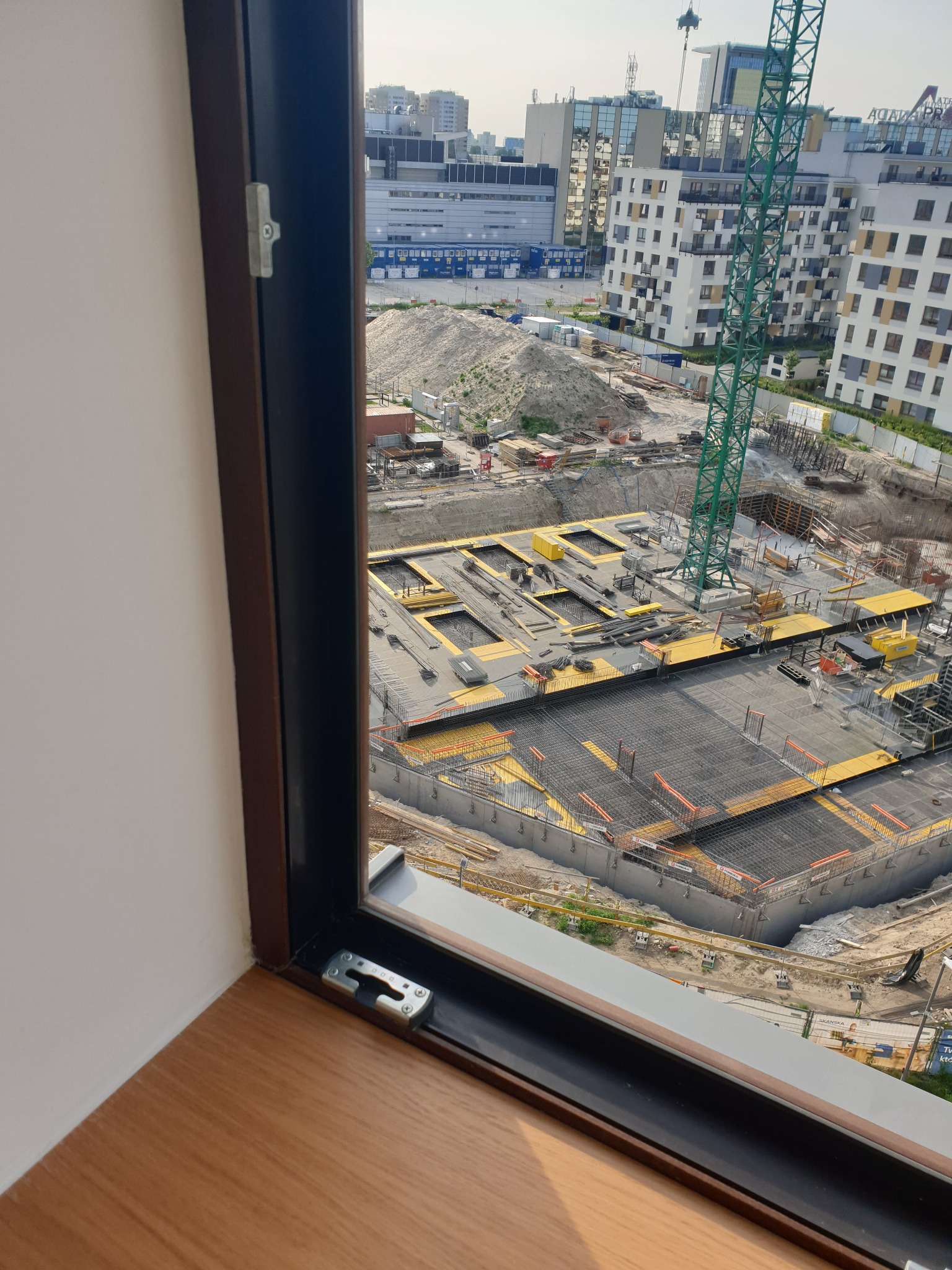Widok z okna na plac budowy z żurawiem, widoczne zbrojenia i fundamenty w żółtym szalunku, w tle zabudowa miejska.