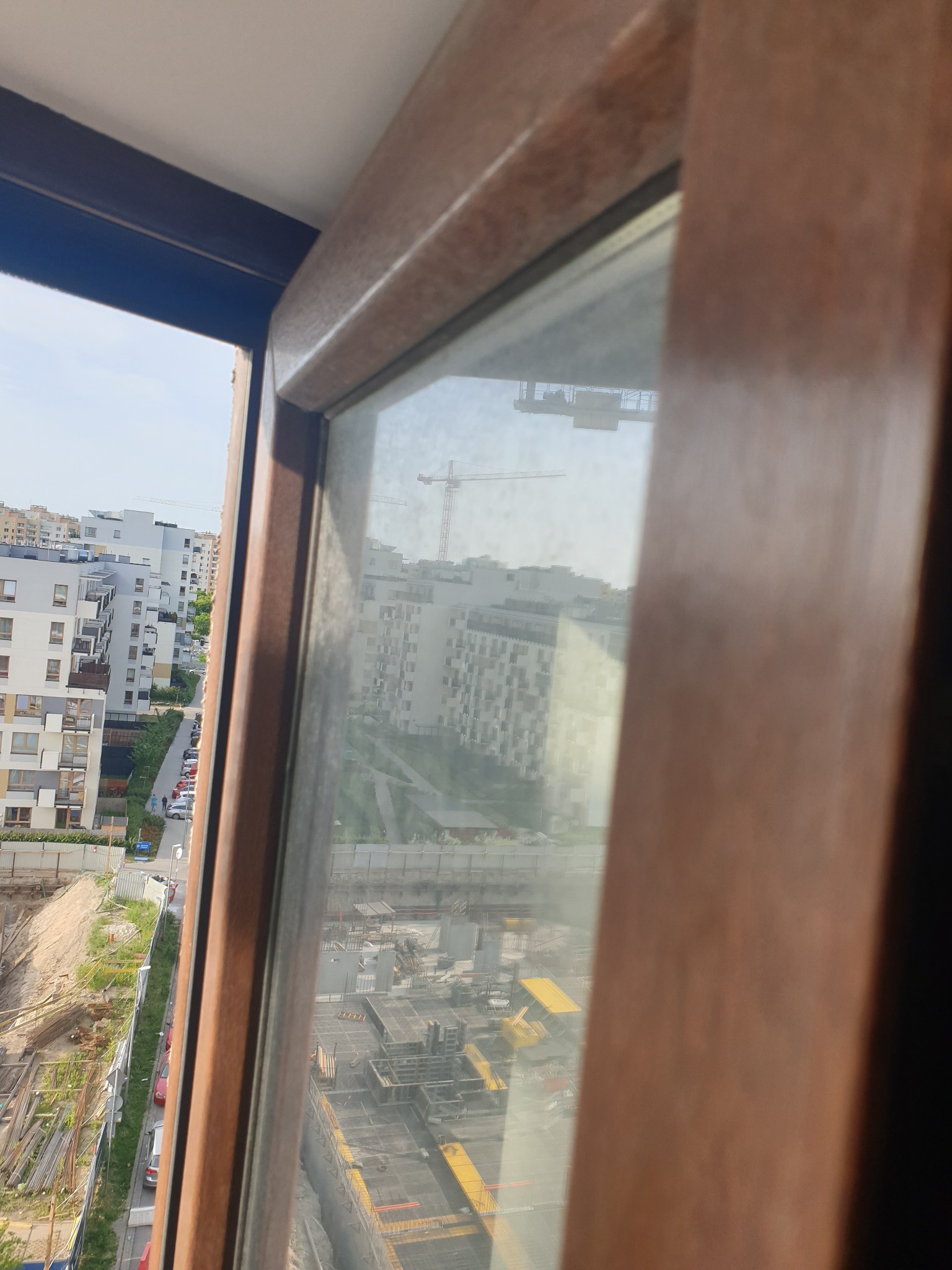 Widok z brudnego okna na osiedle mieszkaniowe i plac budowy z dźwigiem w tle.