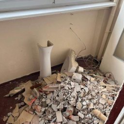 Domin-Trans - Stos gruzu i fragmentów zdemontowanej ceramiki sanitarnej pod ścianą z widocznymi przewodami, po remoncie w pomieszczeniu z oknem i drzwiami.