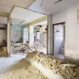 Wnętrze po częściowym wyburzeniu ściany działowej, widoczne gruz i odsłonięte elementy konstrukcyjne, białe ściany z widocznymi śladami po pracach demontażowych.