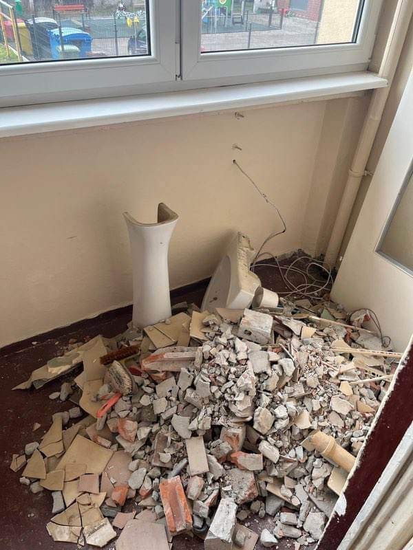 Stos gruzu i fragmentów zdemontowanej ceramiki sanitarnej pod ścianą z widocznymi przewodami, po remoncie w pomieszczeniu z oknem i drzwiami.