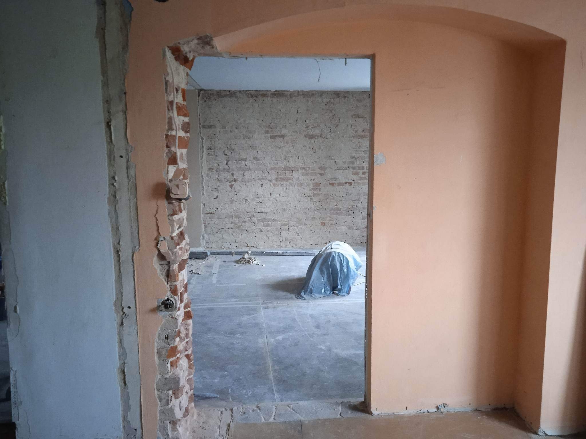 Otwór drzwiowy w trakcie demontażu, odsłonięta cegła, widoczne przewody elektryczne, podłoga zabezpieczona folią, ślady po kuciu na ścianach.