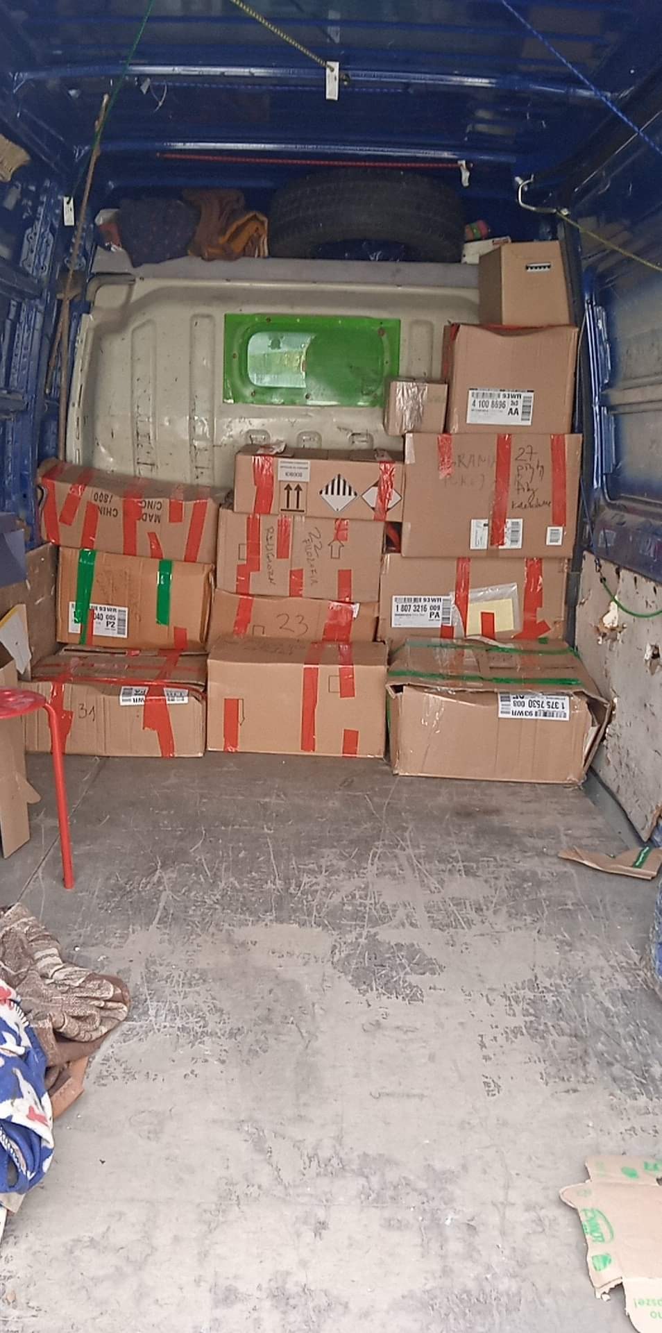 Wnętrze niebieskiego busa wypełnione kartonami różnej wielkości, zabezpieczonymi czerwoną taśmą. Część kartonów zawiera napisy ostrzegawcze i symbole.