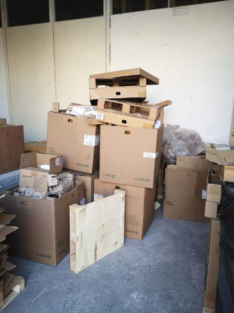 Stos kartonowych pudeł, drewnianych palet i opakowań w magazynie, widoczne ślady użytkowania na podłodze.