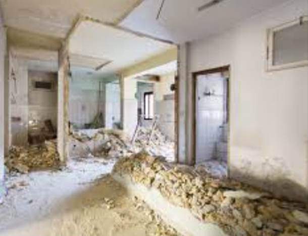 Wnętrze po częściowym wyburzeniu ściany działowej, widoczne gruz i odsłonięte elementy konstrukcyjne, białe ściany z widocznymi śladami po pracach demontażowych.