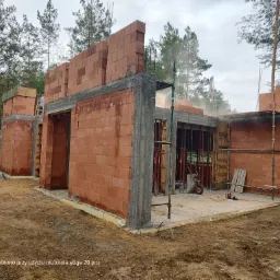 Wznoszenie ścian parterowego budynku z czerwonej cegły ceramicznej, widoczne szalunki betonowe, teren budowy w otoczeniu drzew.