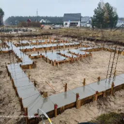 Zalane betonem szalunki fundamentowe na piaszczystym gruncie, widoczne wystające pręty zbrojeniowe i zabudowa jednorodzinna w tle, pochmurny dzień.