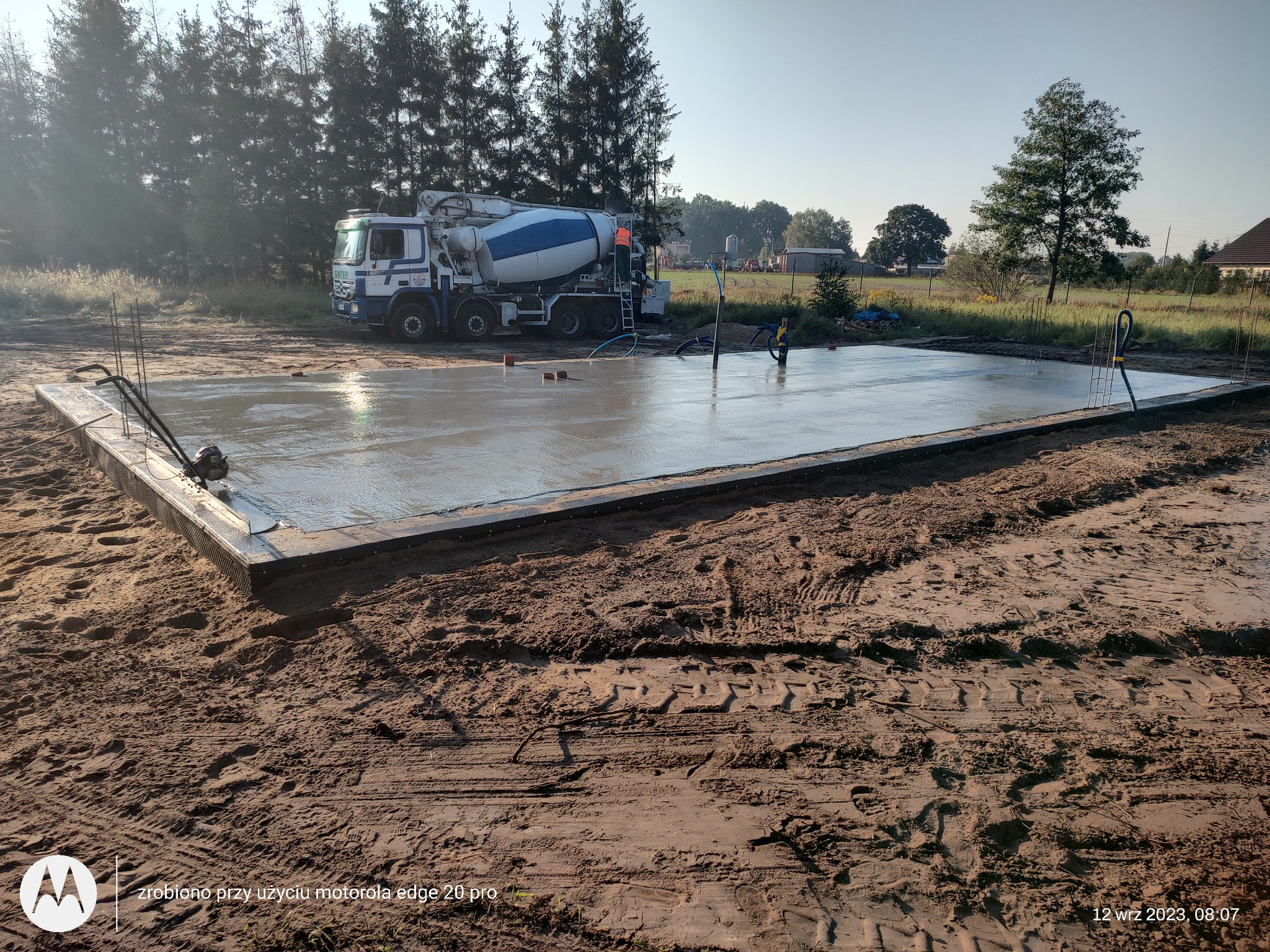 Świeżo wylany fundament pod budynek z widocznym betonowozem w tle, teren budowy z ubitym piaskiem i śladami opon.