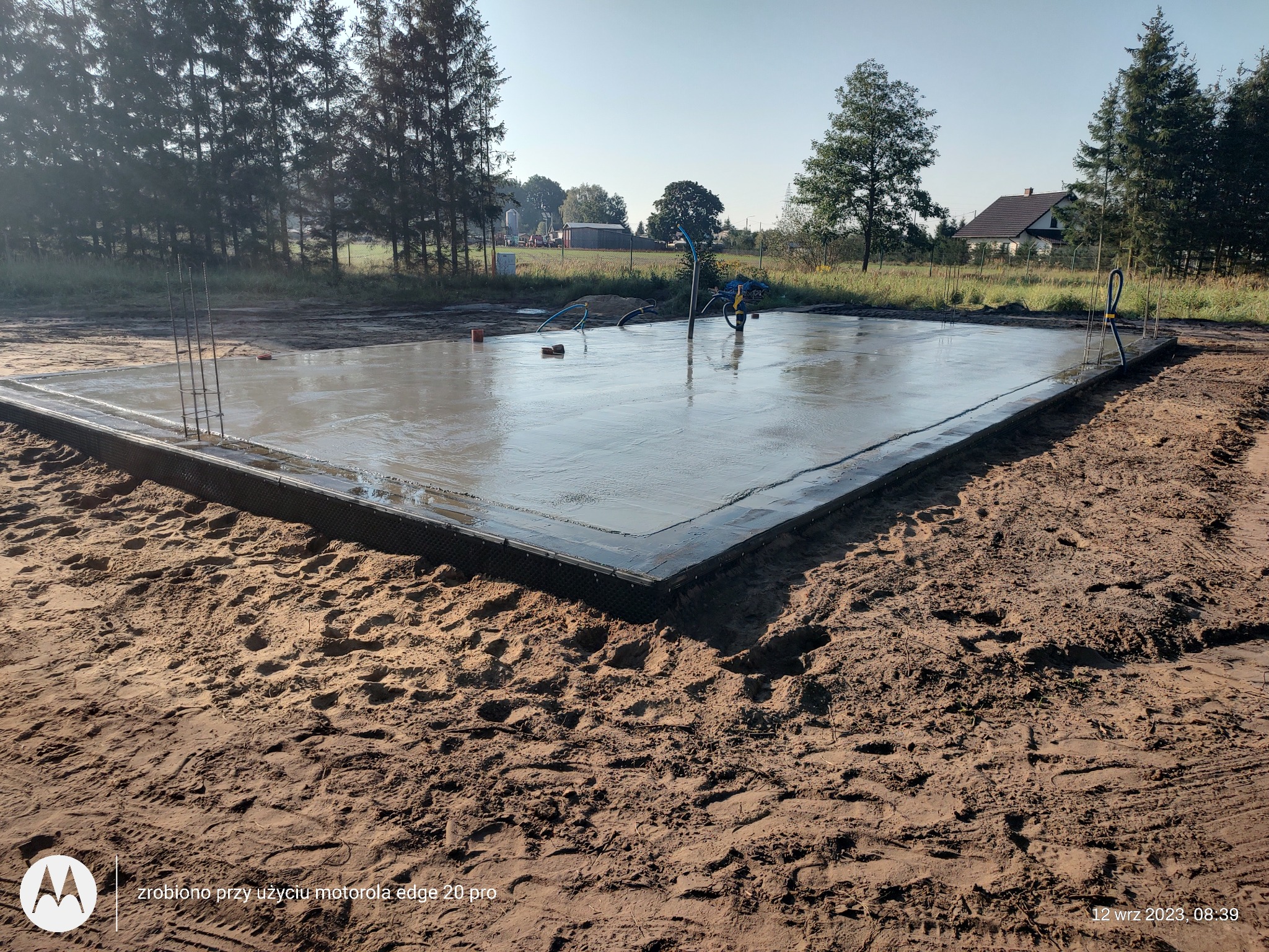 Wylany fundament pod dom jednorodzinny z wystającymi prętami zbrojeniowymi i widocznymi śladami po narzędziach do rozprowadzania betonu na tle drzew i domu.