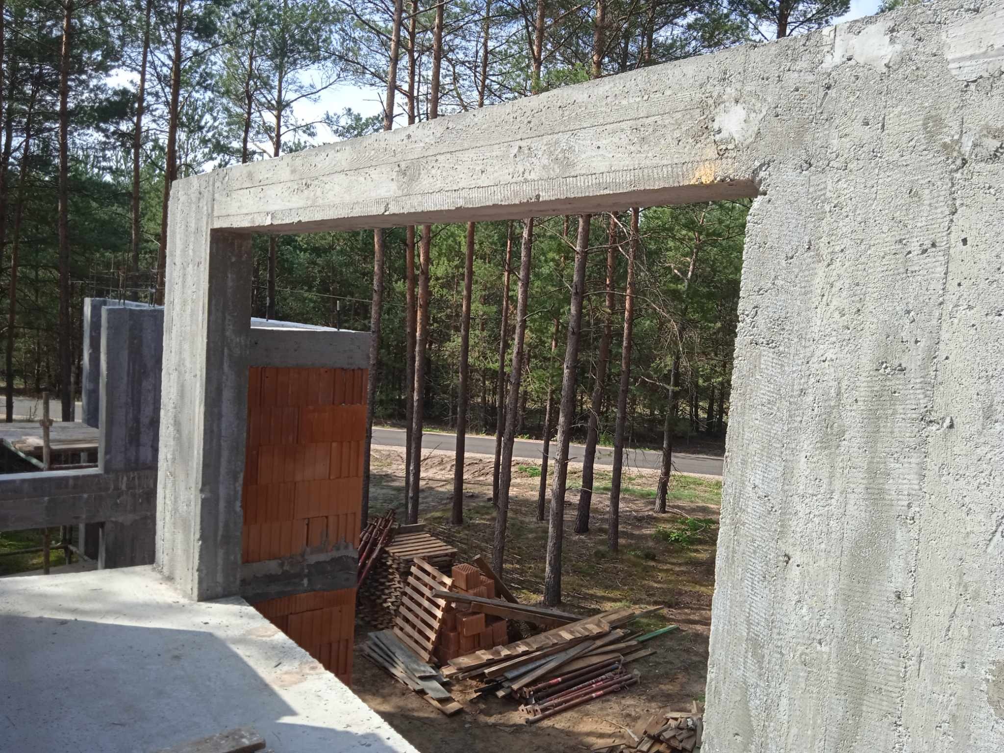 Surowy betonowy szkielet powstającego budynku z widocznymi fragmentami muru z cegły, drewnianymi paletami i podporami budowlanymi w otoczeniu sosnowego lasu.
