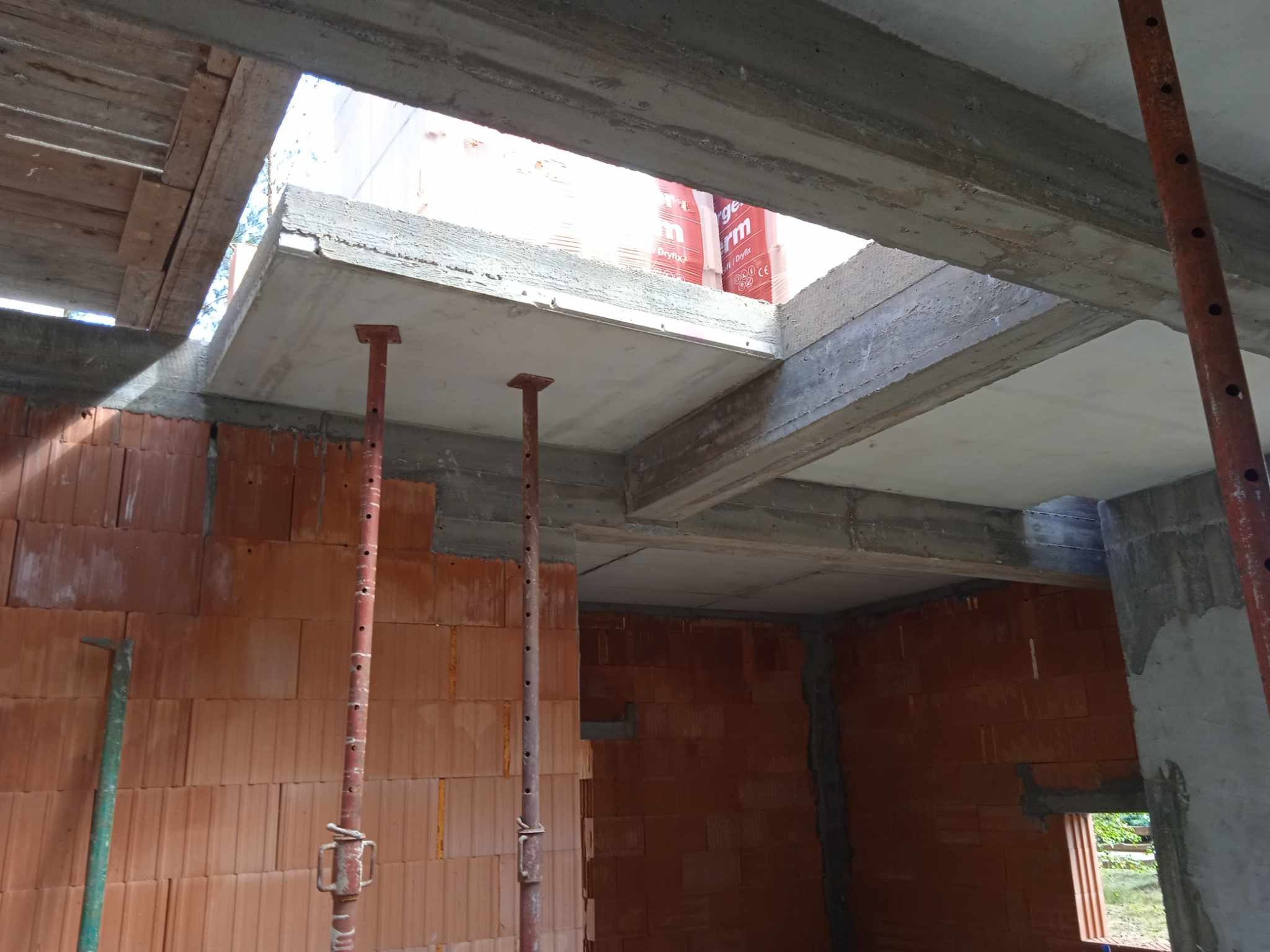 Wnętrze w trakcie budowy: wznoszone ściany z cegły, betonowy strop podparty metalowymi stemplami budowlanymi, widoczne materiały izolacyjne przez otwór w stropie.