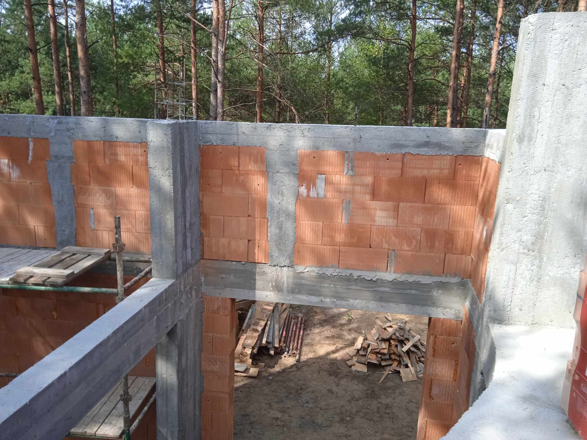 Wznoszenie ścian budynku z pustaków ceramicznych, zbrojone betonowe słupy i nadproża, widoczne drewniane deski i resztki materiałów budowlanych, w tle las.