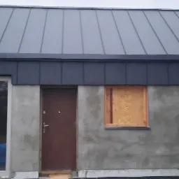 Nowy, grafitowy dach z blachy na surowym budynku w trakcie budowy, z deską zasłaniającą otwór okienny.