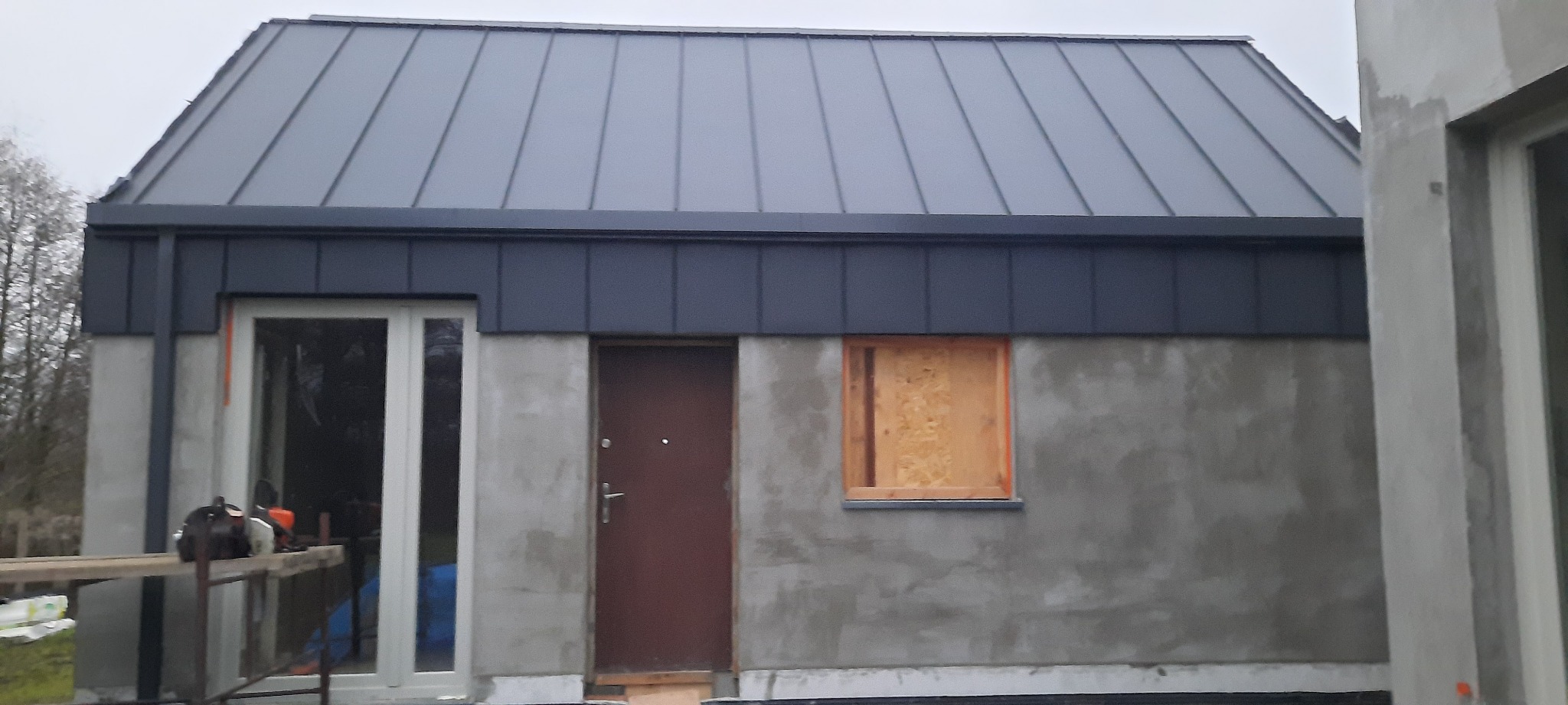 Nowy, grafitowy dach z blachy na surowym budynku w trakcie budowy, z deską zasłaniającą otwór okienny.