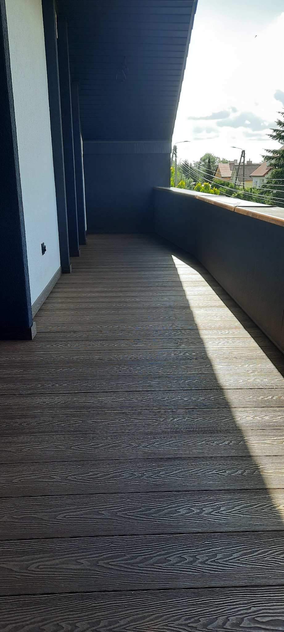Zadaszony balkon z deskami kompozytowymi w odcieniach szarości, z widocznym fragmentem balustrady i słupami nośnymi, oświetlony silnym promieniem słonecznym.