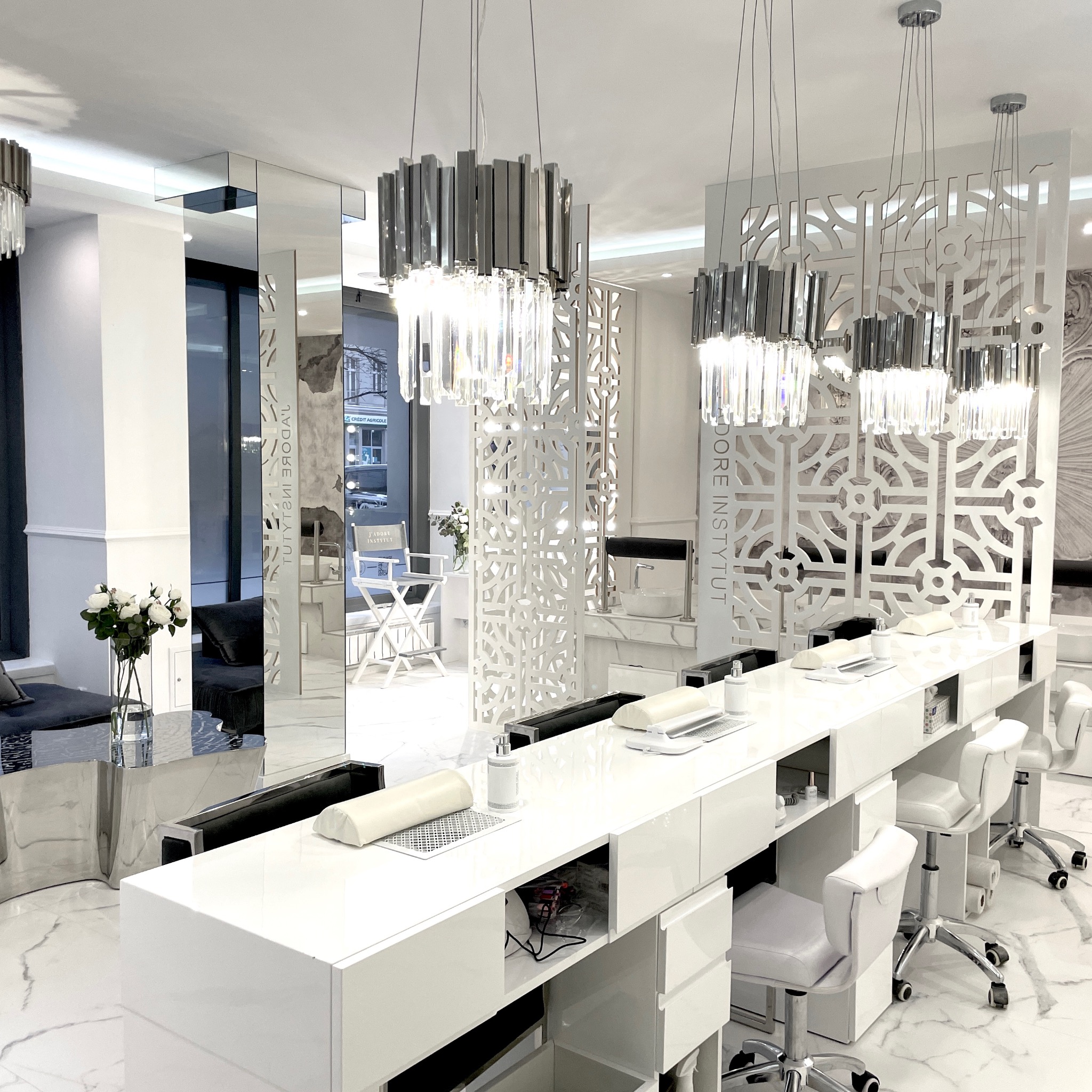 Jasne wnętrze gabinetu z rzędem stanowisk do manicure, nad którymi wiszą kryształowe żyrandole. Białe blaty, krzesła na kółkach i marmurowa podłoga tworzą elegancką przestrzeń.