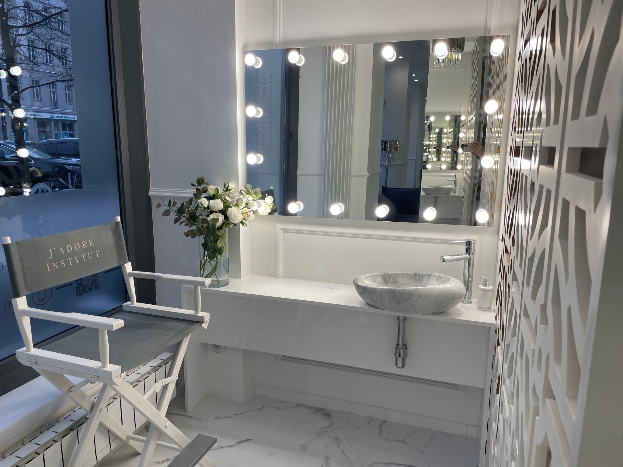 Wnętrze gabinetu z marmurową umywalką i lustrem otoczonym żarówkami, obok krzesło reżyserskie z napisem 'J'ADORE INSTYTUT' i dekoracyjna ścianka działowa.