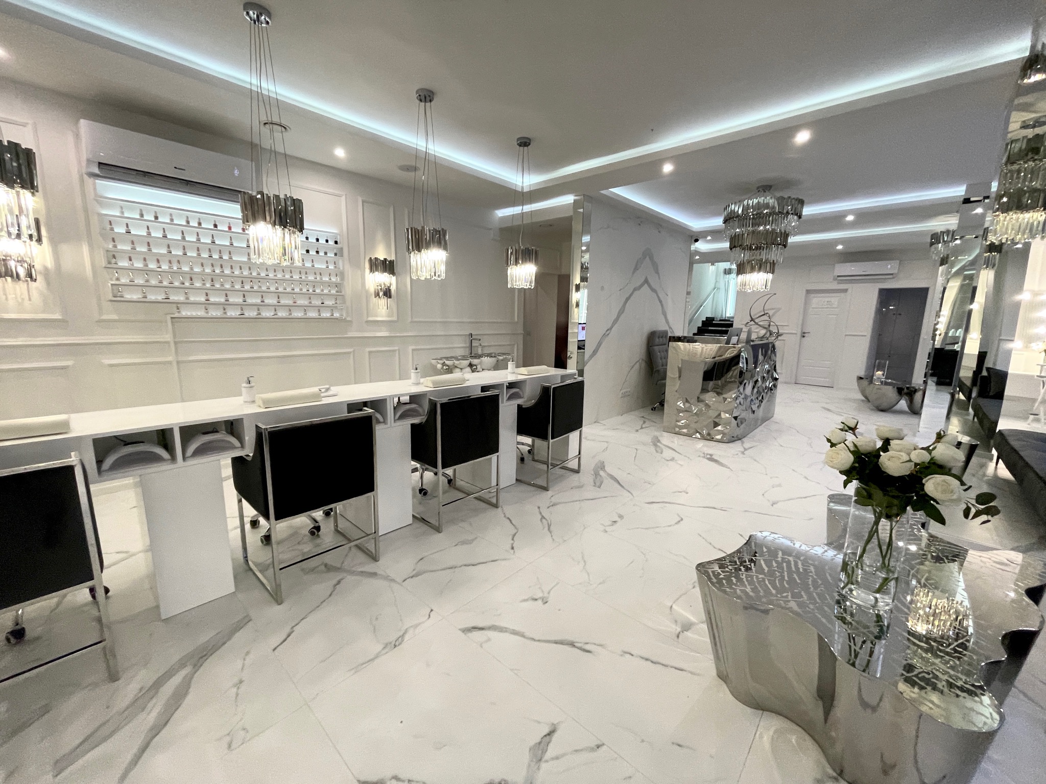 Jasne, przestronne wnętrze salonu kosmetycznego z marmurową podłogą, stanowiskami do manicure z czarnymi krzesłami, ścianą z lakierami do paznokci, nowoczesnymi lampami wiszącymi i dekoracyjnym...
