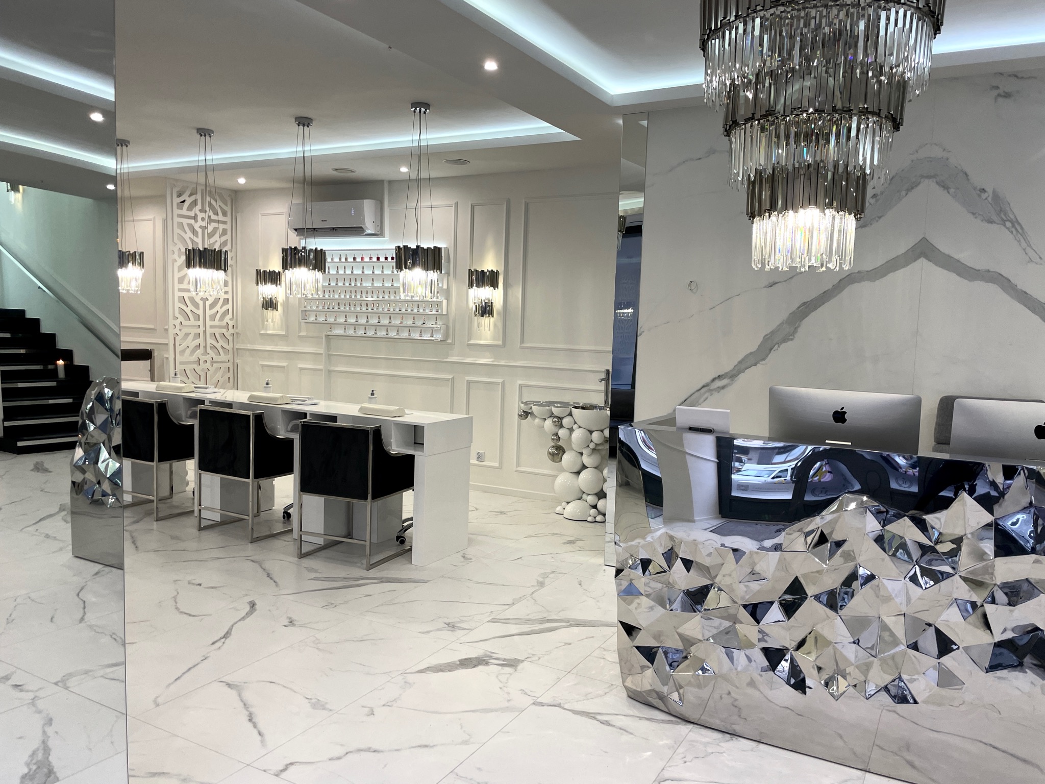 Jasne wnętrze salonu z białymi stanowiskami do manicure, czarnymi krzesłami, ścianą z lakierami do paznokci i designerską ladą recepcyjną z lustrzanymi elementami.