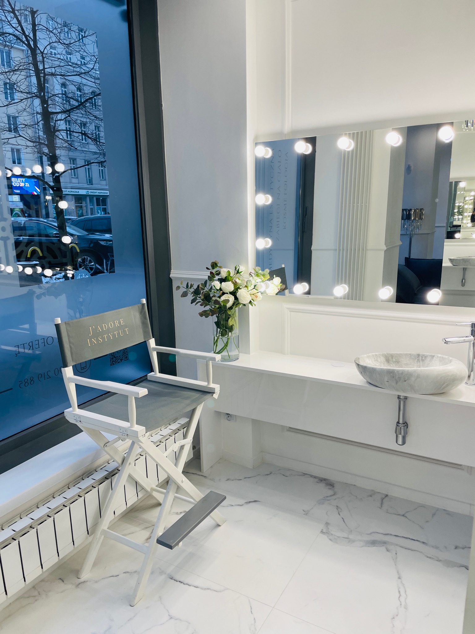 Wnętrze gabinetu kosmetycznego z marmurową umywalką, lustrem z oświetleniem, bukietem białych kwiatów w wazonie i krzesłem reżyserskim z napisem 'J'ADORE INSTYTUT' na tle okna z widokiem na ulicę.