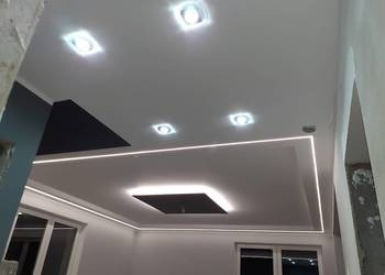 Nowoczesny sufit podwieszany z oświetleniem LED w kształcie prostokąta, z dodatkowymi punktowymi lampami sufitowymi. Widoczne fragmenty ścian pomalowanych na biało i granatowo oraz okna. W tle...