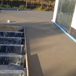 Świeżo wylana, gładka posadzka na tarasie z widocznymi zabezpieczonymi schodami i drzewami w tle, w słoneczny dzień.