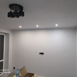 Szare wnętrze pokoju z zamontowanym oświetleniem sufitowym: czarna lampa wisząca i halogeny, widoczna instalacja elektryczna w ścianie, butelka wody i karton na podłodze.