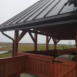 Zadaszona drewniana altana z metalowym dachem w kolorze ciemnej czekolady, widok na rozległe łąki w pochmurny dzień.