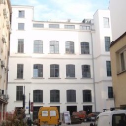 InvestComplex-2 - Biały budynek wielopiętrowy z ciemnymi oknami w otoczeniu innych budynków, widoczny samochód dostawczy i materiały budowlane na placu